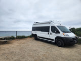 White **RV rental** van parked on a dirt lot overlooking the ocean, with a picnic table and cloudy sky.