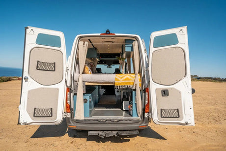 RV rental: A white camper van with rear doors open, revealing a cozy custom interior with a bed, parked on a dirt road by the ocean.