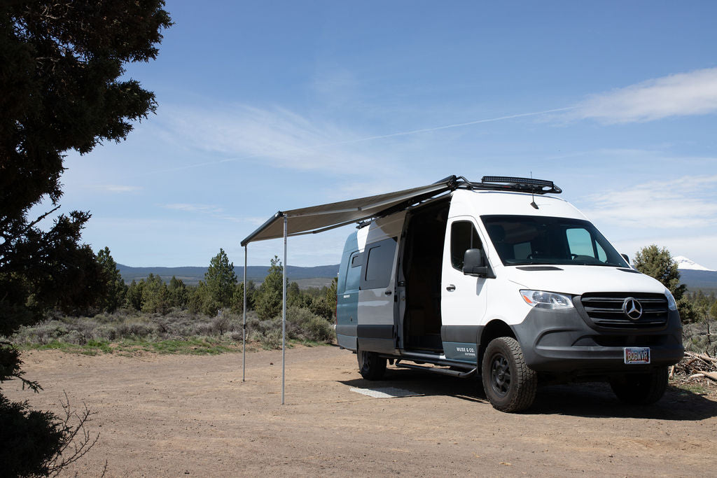 White and teal camper van RV rental with awning extended, parked on a dirt track amidst trees for outdoor adventures.