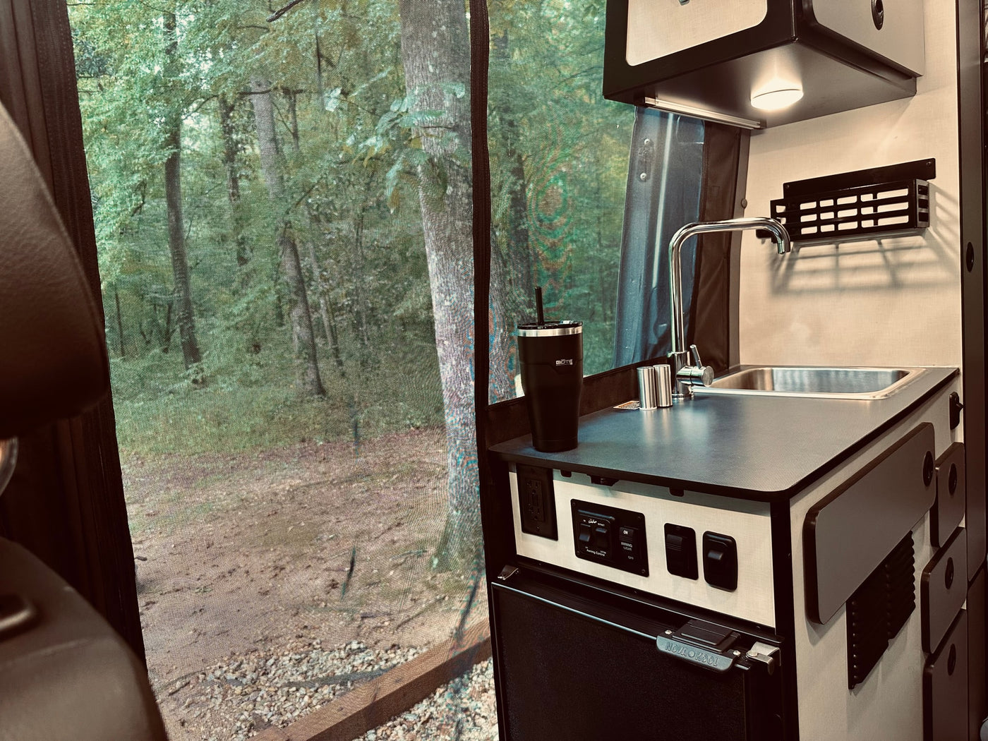 Inside an RV rental: a kitchenette with sink, controls, and fridge, looking out to a serene forest scene through a screen. Perfect for camping! — photo 6