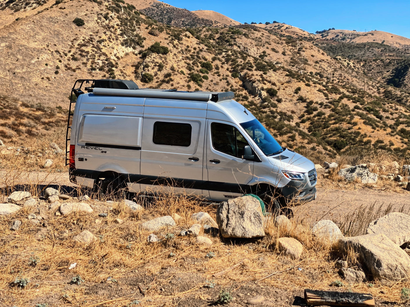 Silver Winnebago Revel 4x4 camper van, ideal for RV rental adventures, parked on a dry mountain trail with rocks and sparse vegetation.