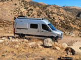 Silver Winnebago Revel 4x4 camper van, ideal for RV rental adventures, parked on a dry mountain trail with rocks and sparse vegetation.