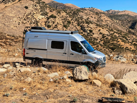 Silver Winnebago Revel 4x4 camper van, ideal for RV rental adventures, parked on a dry mountain trail with rocks and sparse vegetation.