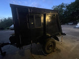 A dark, enclosed custom camper or utility trailer with a window & door, parked outdoors. Possibly for an RV rental service.
