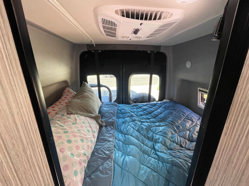 Inside this **rv rental**, a compact bed with colorful bedding, overhead AC unit, and rear windows offer a cozy travel experience. — photo 6