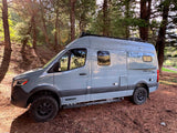 A grey Winnebago Revel 4x4 camper van, ideal for your next rv rental, sits in a sunny pine forest, ready for adventure.
