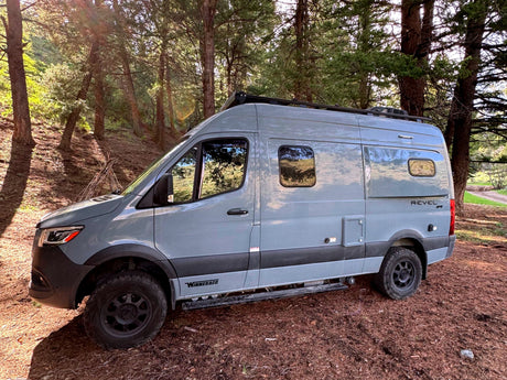 A grey Winnebago Revel 4x4 camper van, ideal for your next rv rental, sits in a sunny pine forest, ready for adventure.