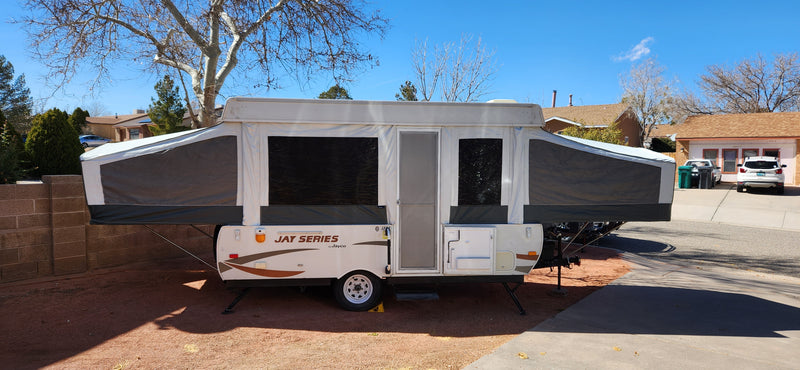 A white and gray pop-up camper, fully expanded, is parked in a yard, ideal for an RV rental. Houses are visible in the sunny background. — photo 1