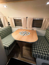 Inside a cozy RV rental: green plaid dinette seating, wooden table with compass design, and windows with curtains.