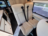 RV rental interior: two light-colored seats with seatbelts, a table, kitchen with microwave, hex-pattern flooring.