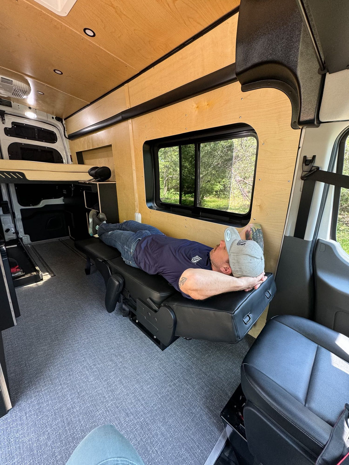  a man relaxes in a custom camper van with a cozy bed & kitchen.|Enjoying a meal inside a custom RV rental. Features a clever interior kitchen with fold-out tables & water tanks for mobile living.|Inside an RV rental: Man relaxes on a modular seat in this custom camper van conversion.|A man with tattoos enjoys a sandwich and chips inside a cozy RV rental — photo 7