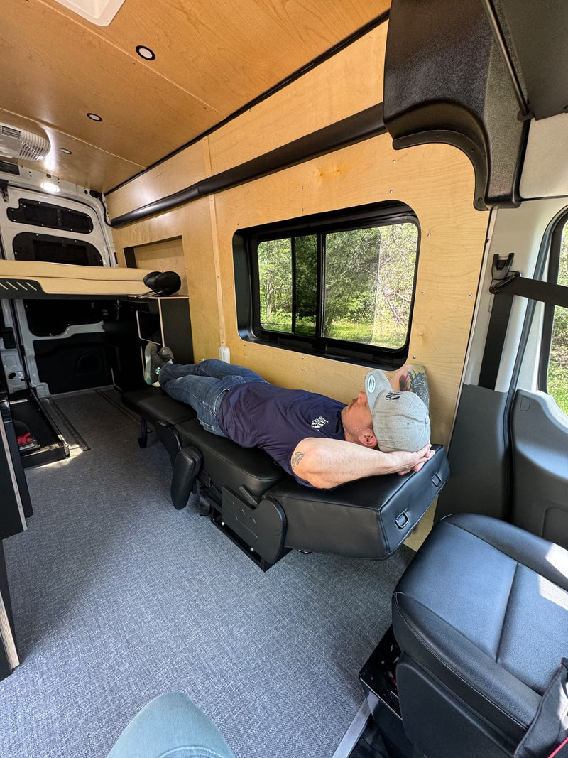 a man relaxes in a custom camper van with a cozy bed & kitchen.|Enjoying a meal inside a custom RV rental. Features a clever interior kitchen with fold-out tables & water tanks for mobile living.|Inside an RV rental: Man relaxes on a modular seat in this custom camper van conversion.|A man with tattoos enjoys a sandwich and chips inside a cozy RV rental — photo 7