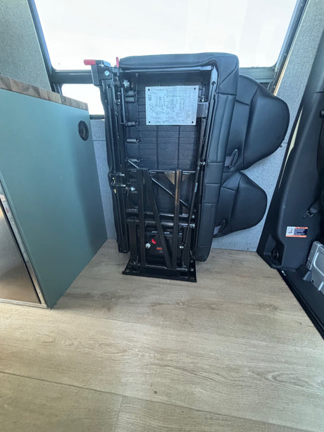 An RV rental interior features a black foldable seat/bed mechanism stowed, next to teal cabinets on light wood-look flooring.
