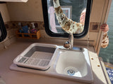 Inside an RV rental: A kitchen sink with a dish drying rack, cleaning supplies, and a person cleaning the window from outside.