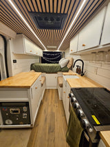 This rv rental features a cozy bed, well-equipped kitchen with a stove and sink, and modern wood-slat ceiling design.