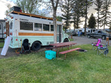 Custom converted 'Skoolchella' bus in an RV campground setup with picnic gear. Explore RV rental options for travel!