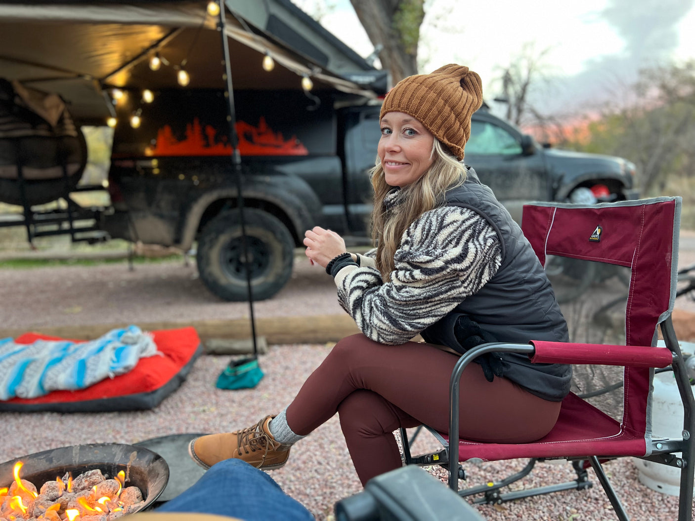 Cozy RV rental evening. A woman smiles by a fire pit; truck with awning & lights in back. Perfect for outdoor relaxation.