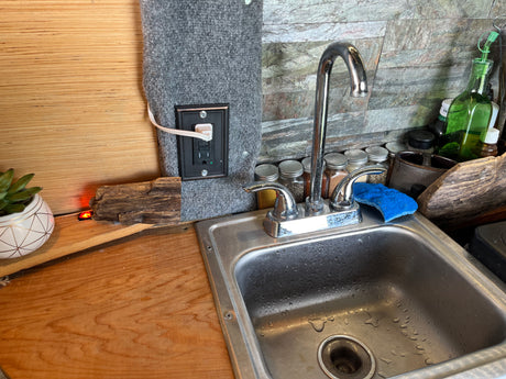 RV rental's compact kitchen setup: stainless sink, chrome faucet, spice jars, and a power outlet on carpeted wall.