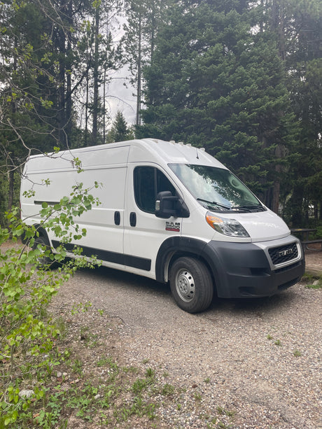 A white Ram ProMaster van is parked on a gravel campsite amidst trees, perfect for your next rv rental adventure.