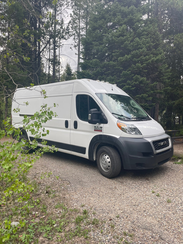 A white Ram ProMaster van is parked on a gravel campsite amidst trees, perfect for your next rv rental adventure.