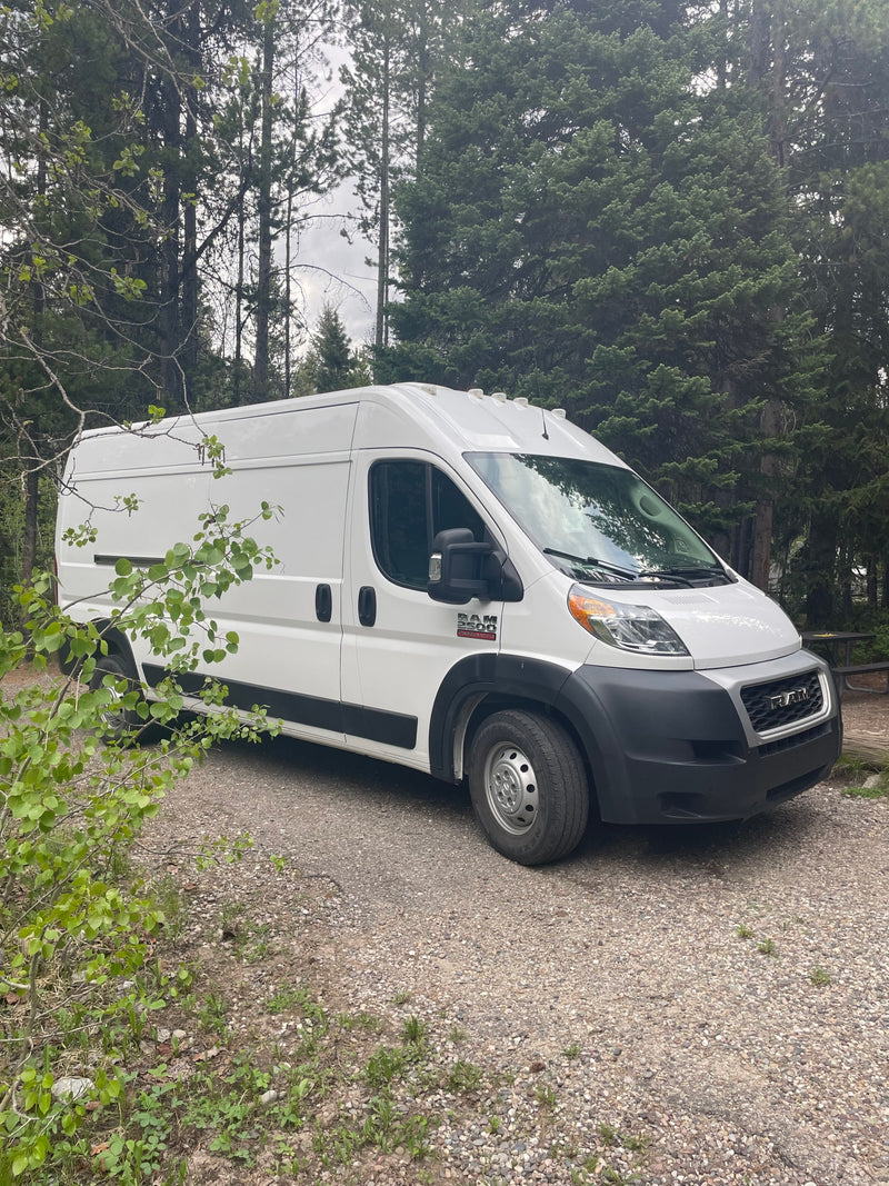 A white Ram ProMaster van is parked on a gravel campsite amidst trees, perfect for your next rv rental adventure. — photo 1