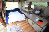 RV rental interior: cozy bed, compact kitchen with stove, sink, mugs, and a scenic outdoor view.