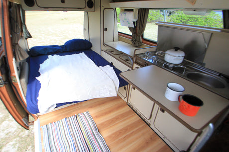 RV rental interior: cozy bed, compact kitchen with stove, sink, mugs, and a scenic outdoor view.