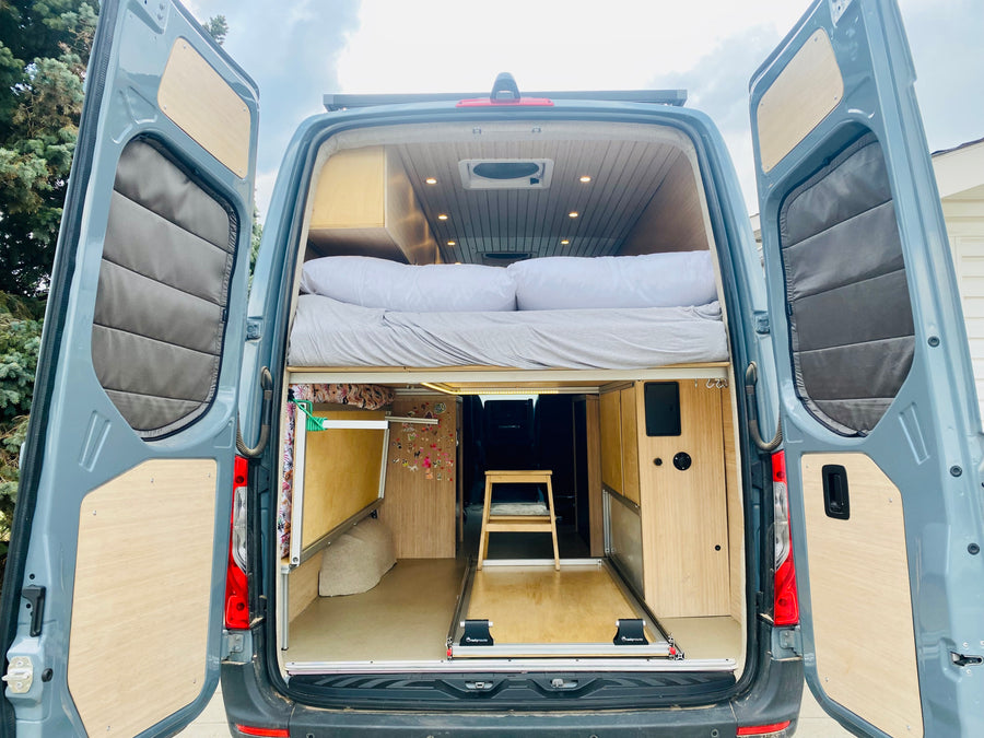 Rear view of a spacious camper van interior featuring a cozy upper bed, pull-out storage, and wood finishes, ideal for an RV rental.