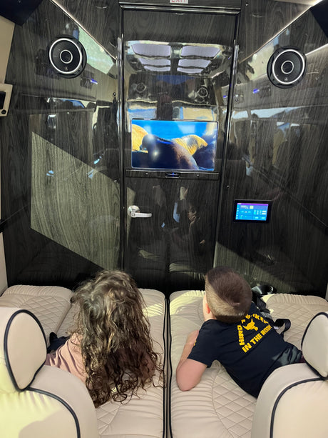 Kids enjoy a movie on the screen in a luxury RV rental, with comfortable seating and a sleek wood-grain interior.