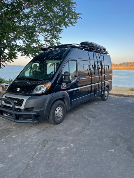 Black Ram ProMaster 2500 RV rental parked lakeside, equipped with a roof rack & bull bar, perfect for outdoor travel.