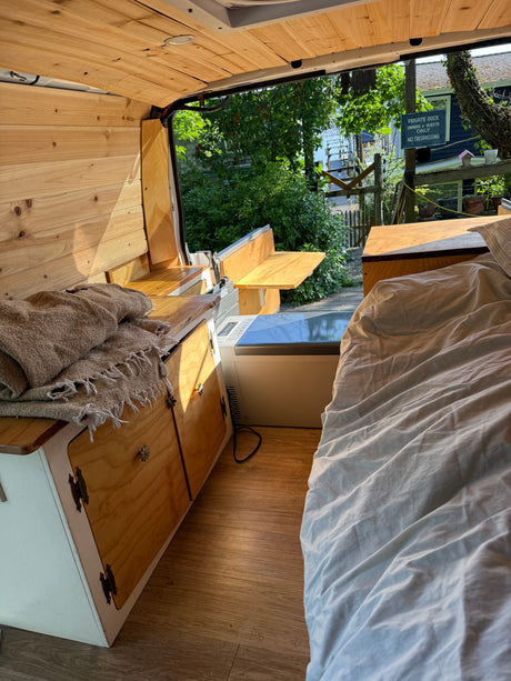 Cozy RV rental interior: wooden walls, a comfy bed, kitchenette, and portable fridge, offering a serene outdoor view.