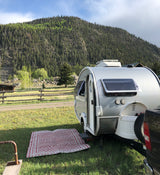 RV rental: A white teardrop camper on grass with an outdoor rug, against forest-covered mountains. Perfect for camping!