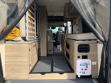 Inside an RV rental: rear view of the living area with light wood cabinetry, flexible bed/seating, and utility panel.