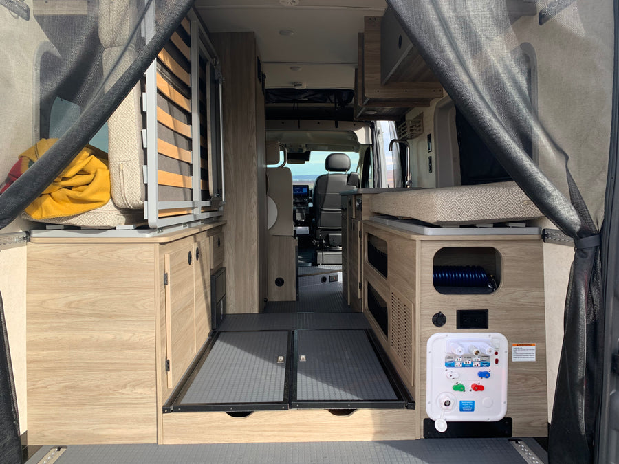 Inside an RV rental: rear view of the living area with light wood cabinetry, flexible bed/seating, and utility panel.