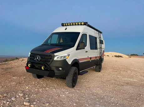 A rugged white Mercedes Sprinter adventure van, equipped for off-road travel, parked on a dirt road. Great for an RV rental.