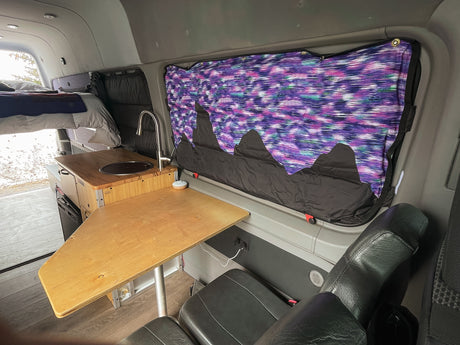 Inside an RV rental camper van: elevated bed, sink kitchen, and a wooden dining table with seating. Colorful window covers add flair.