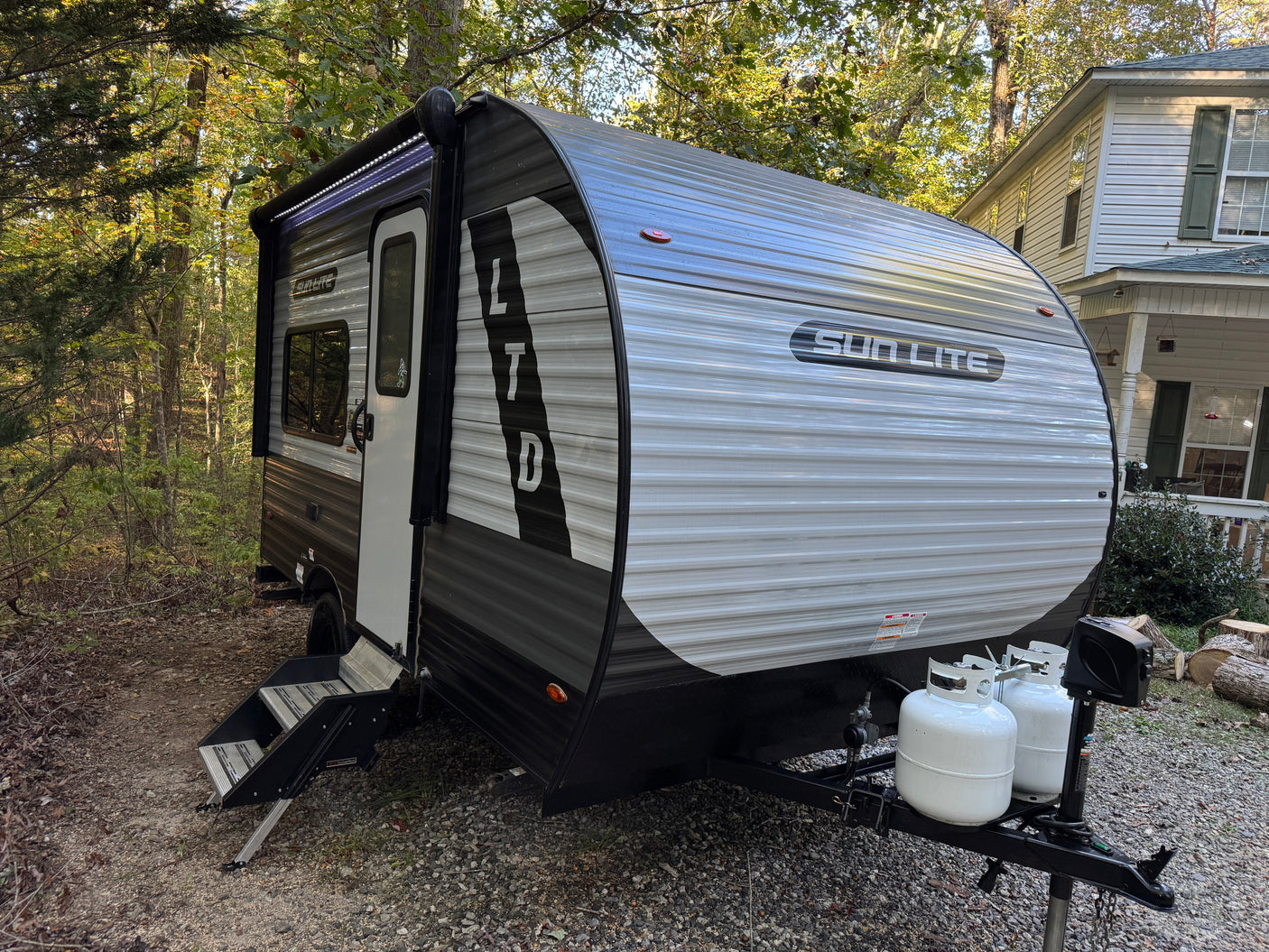 This Sun Lite LTD travel trailer, possibly an RV rental, is parked in a wooded area with its awning extended and propane tanks visible.