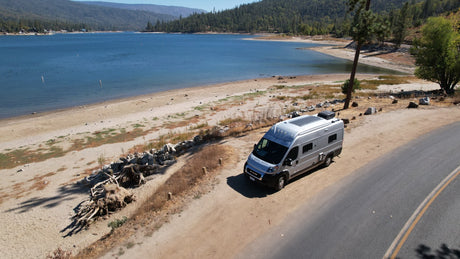 An RV rental offers prime views. A silver camper van is parked on a dirt pull-off overlooking a large lake and sandy beach amidst mountains.