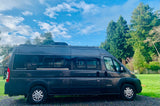 Dark gray Winnebago Travato RV rental, a modern camper van, parked on gravel next to green trees under a blue sky.