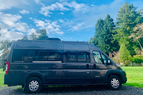Dark gray Winnebago Travato RV rental, a modern camper van, parked on gravel next to green trees under a blue sky.