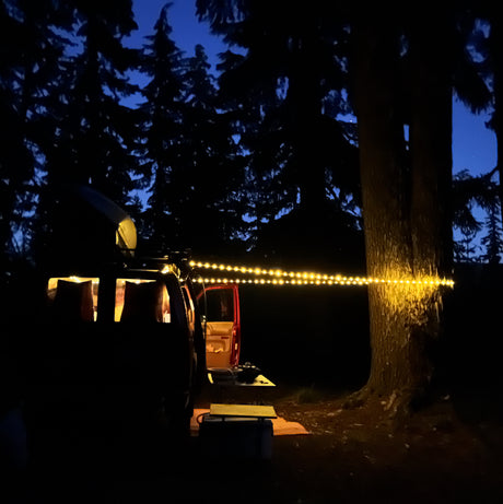 Cozy night setup with an RV rental. String lights illuminate the van's interior and a tree in the dark forest campsite.