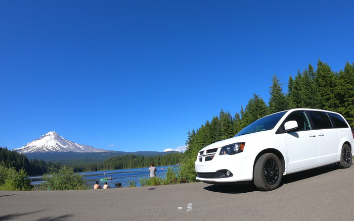 White minivan (RV rental) parked by a scenic lake with snow-capped Mt. Hood. People enjoy water activities & forest views.