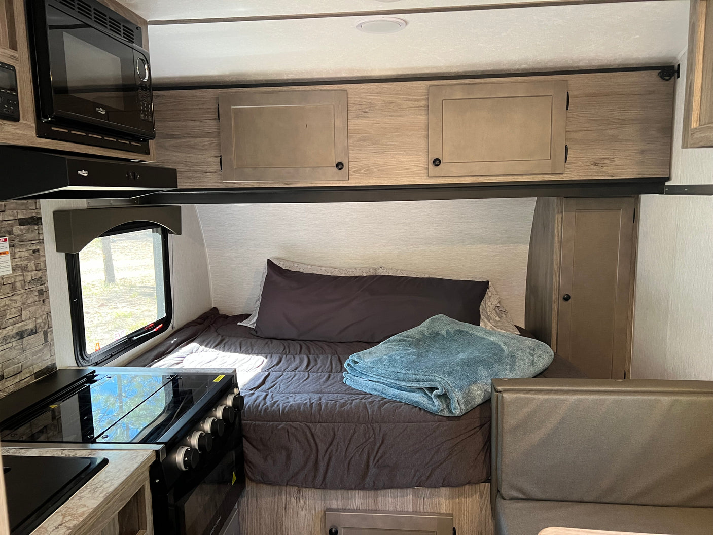 RV rental interior: kitchen with microwave, range, and sink, plus a bed with a blue blanket, overhead cabinets, and dinette seating.
