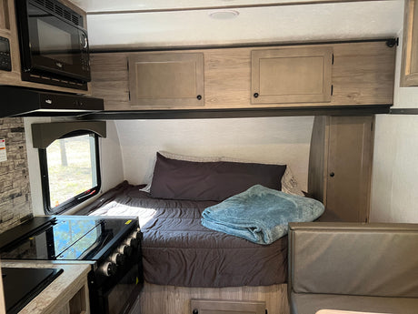RV rental interior: kitchen with microwave, range, and sink, plus a bed with a blue blanket, overhead cabinets, and dinette seating.