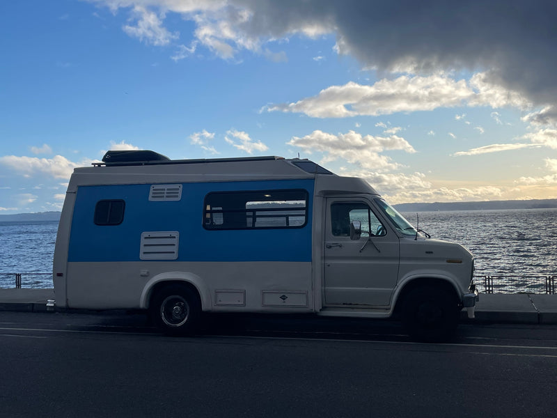 A two-tone camper van RV rental parked by the water, under a blue sky with clouds. Ready for a scenic road trip adventure. — photo 1