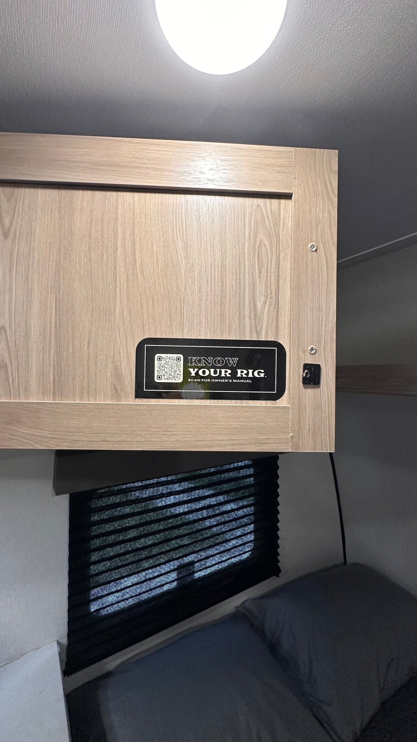 Inside an RV rental: Overhead cabinet with owner's manual QR code, window with blinds, and a comfortable bed.