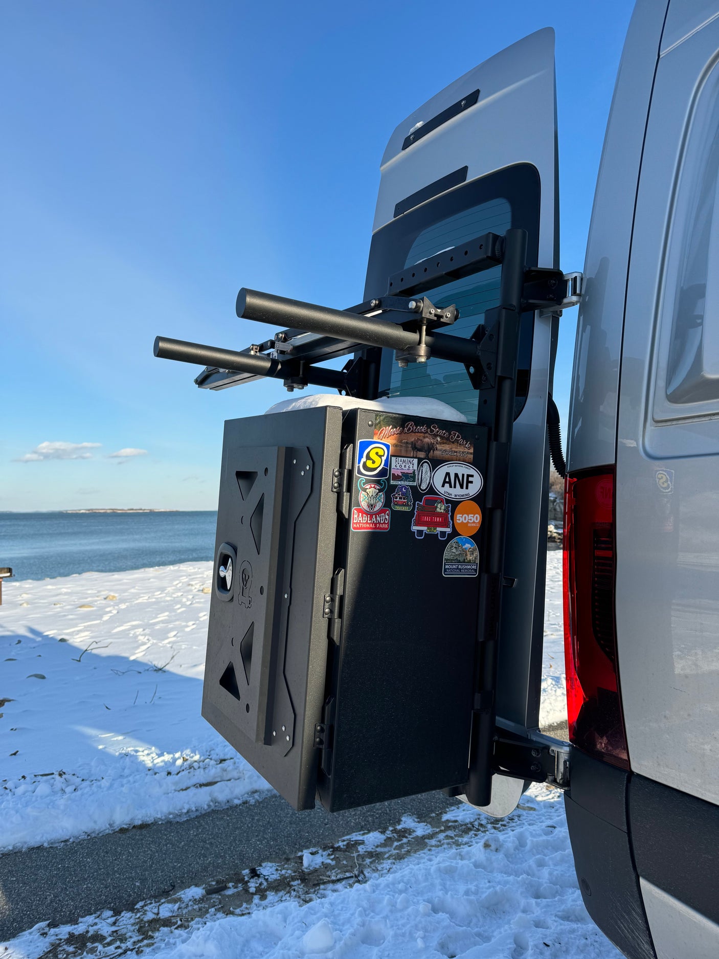 A silver van (likely an **RV rental**) with a black rear cargo box, gear rack, and travel stickers is parked on a snowy shore by the ocean.
