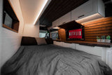 This modern rv rental interior features a comfy bed, flip-down Netflix TV, and sink. White cabinets, wood accents, and bright lighting create a cozy space.