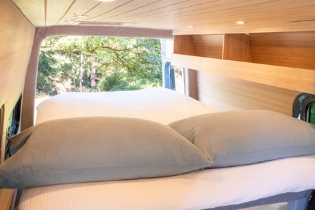 Cozy RV rental bedroom with warm wood interior, comfortable bed, pillows, and a peaceful view of lush trees outside.