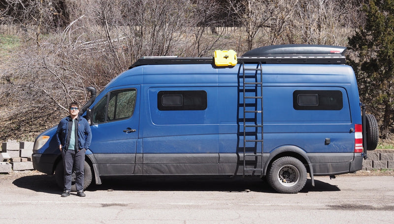 Man stands beside a blue RV rental camper van, featuring a roof rack, cargo box, and ladder, in an outdoor setting. — photo 1